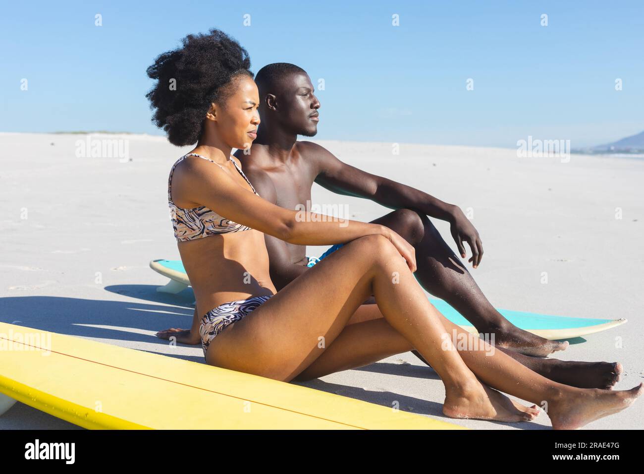 Felice coppia afro-americana seduta sulla spiaggia soleggiata con tavole da surf che guardano al mare Foto Stock