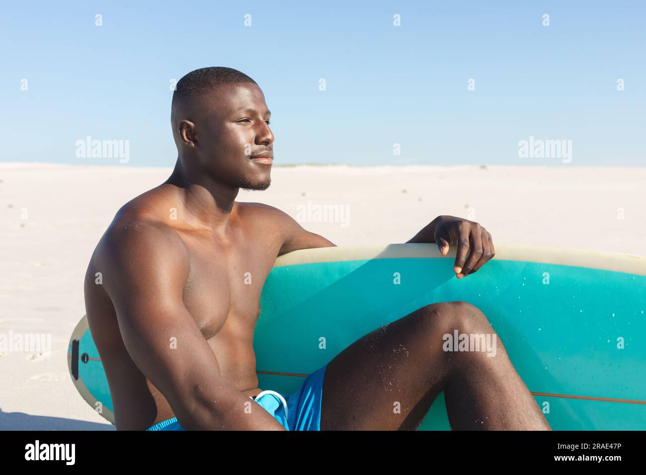 Felice afro-americano seduto sulla spiaggia soleggiata con tavola da surf che guarda al mare Foto Stock