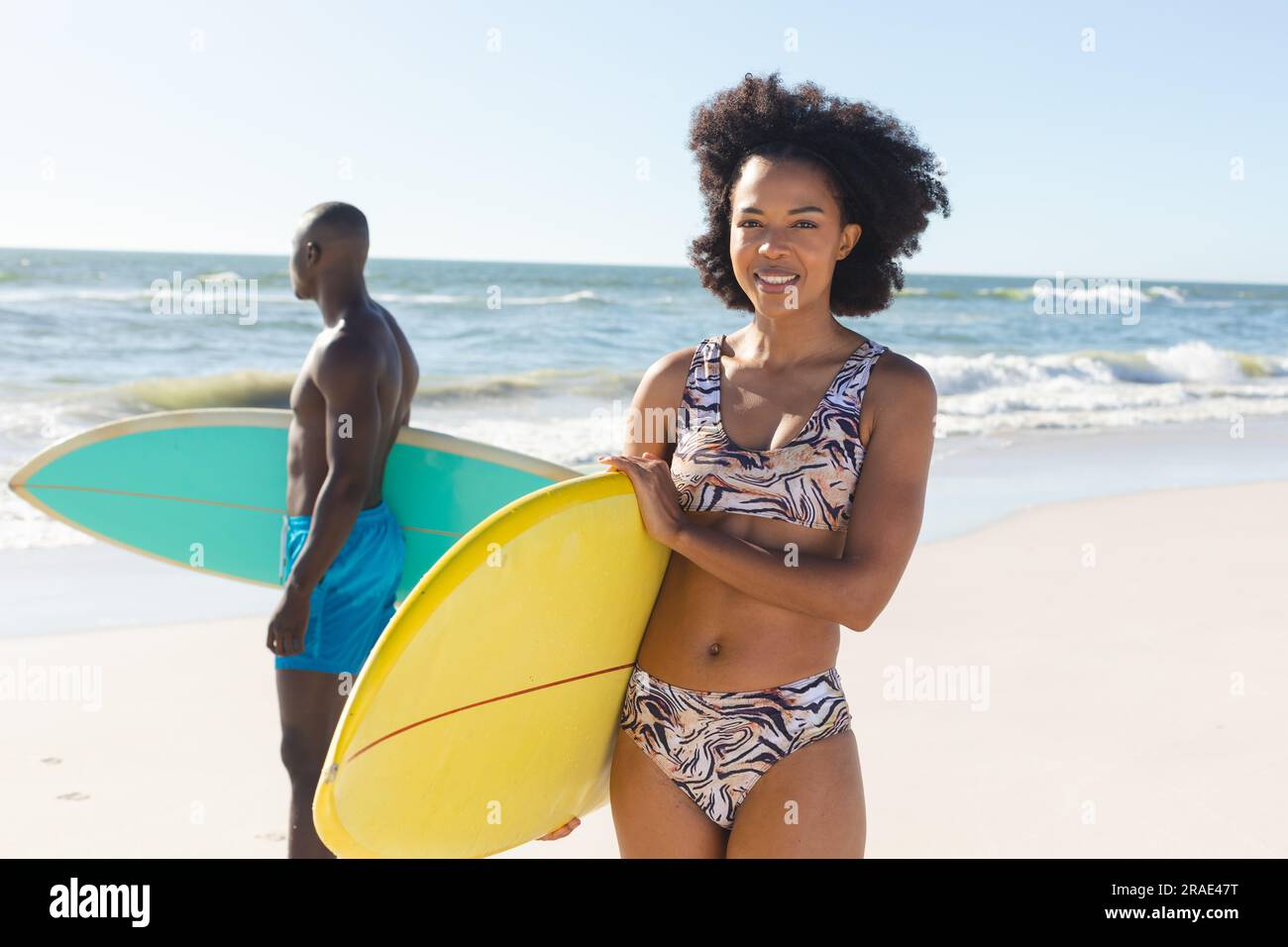 Ritratto di una donna afroamericana felice che tiene una tavola da surf in piedi sulla spiaggia soleggiata con un surfista maschio Foto Stock