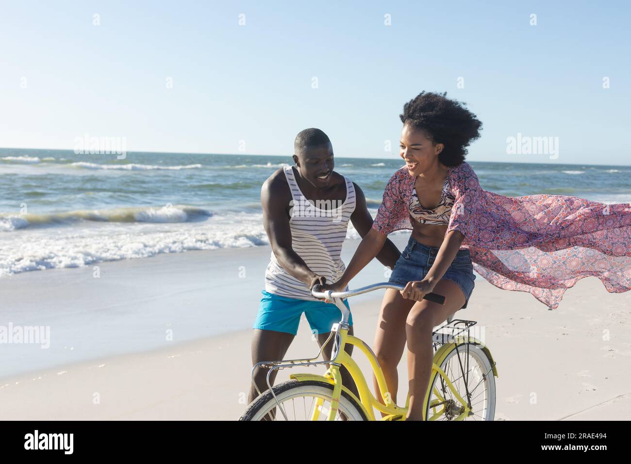 Felice coppia afro-americana che si diverte a camminare e andare in bicicletta sulla spiaggia soleggiata vicino al mare, spazio fotocopie Foto Stock