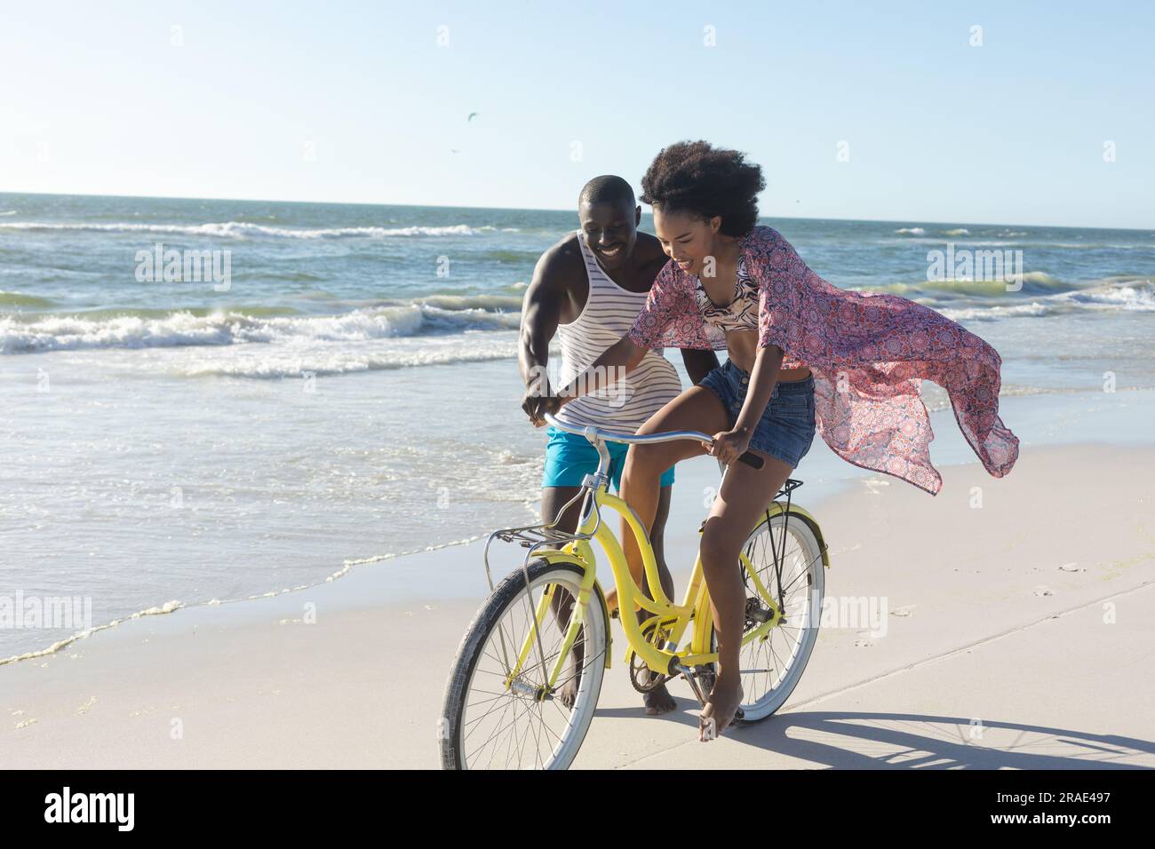 Felice coppia afro-americana che si diverte a camminare e andare in bicicletta sulla spiaggia soleggiata vicino al mare, spazio fotocopie Foto Stock