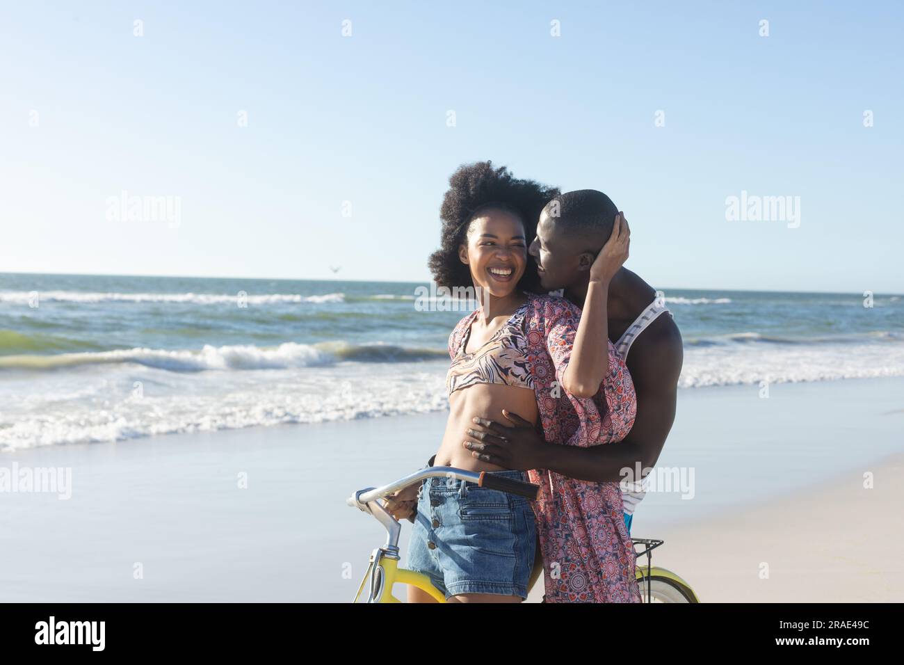 Felice coppia afro-americana che abbraccia la bicicletta sulla spiaggia soleggiata in riva al mare Foto Stock