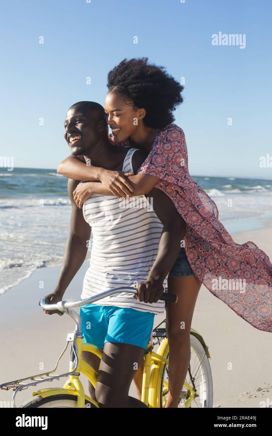 Felice coppia afro-americana che abbraccia la bicicletta sulla spiaggia soleggiata in riva al mare Foto Stock