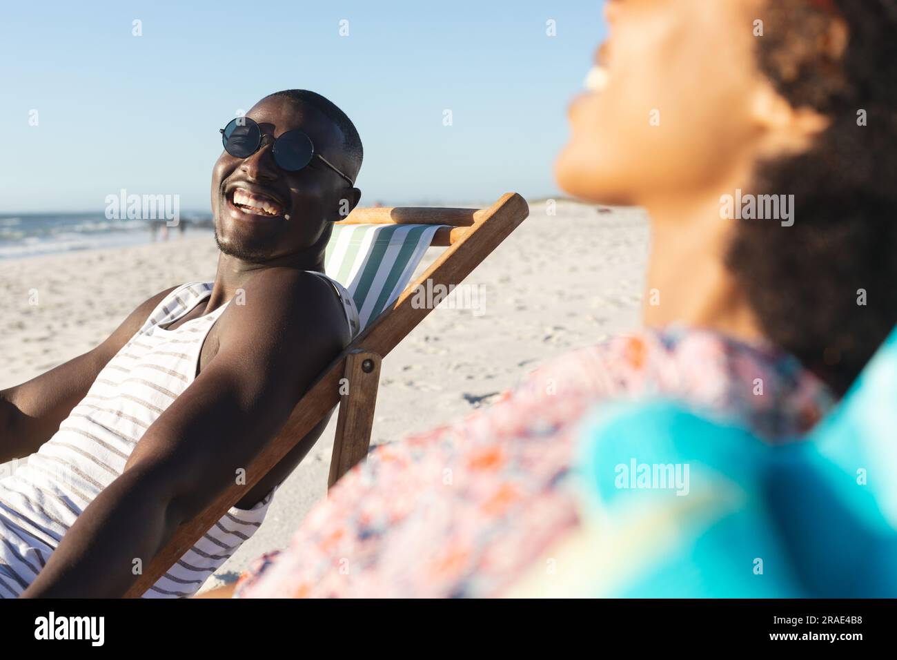 Felice coppia afro-americana in occhiali da sole seduti su sdraio ridendo sulla spiaggia soleggiata Foto Stock
