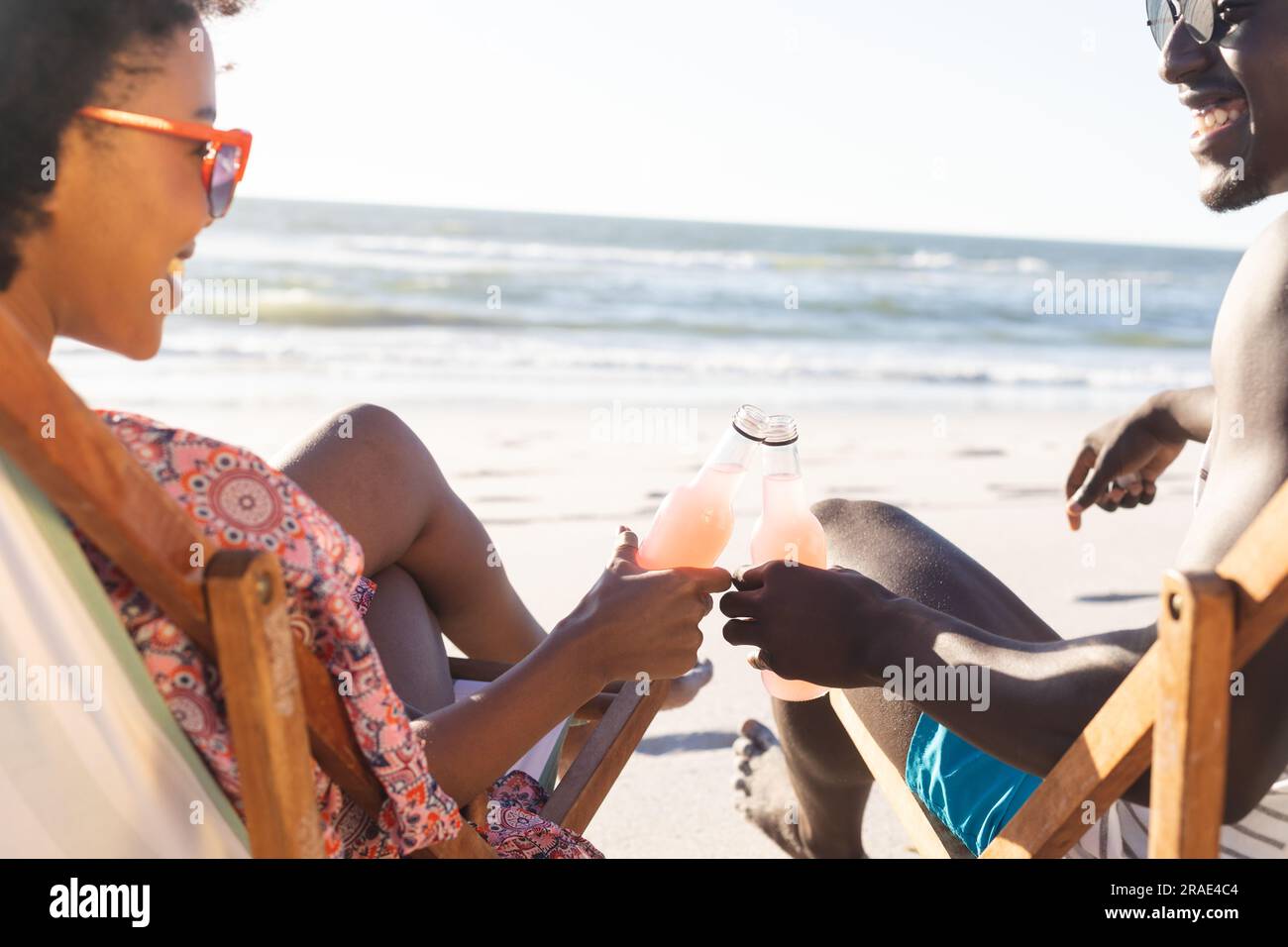 Felice coppia afro-americana seduta su sdraio a fare un brindisi con bottiglie sulla spiaggia soleggiata Foto Stock