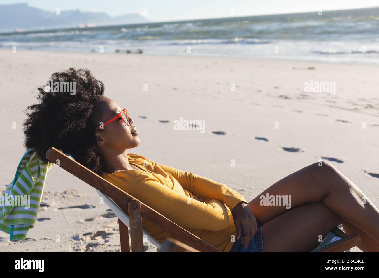 Felice donna afro-americana con occhiali da sole che si rilassa sulla sdraio sulla spiaggia soleggiata in riva al mare Foto Stock