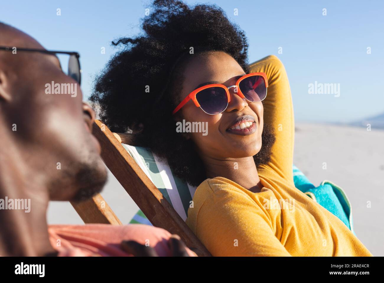 Felice coppia afro-americana con occhiali da sole seduti su sdraio sorridendo sulla spiaggia soleggiata Foto Stock