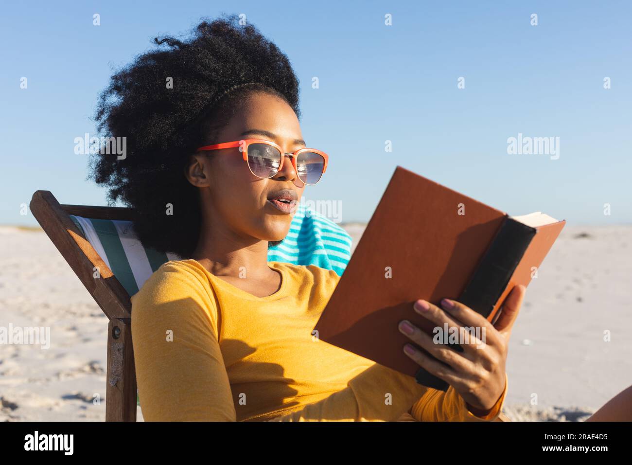 Felice donna afro-americana con occhiali da sole seduta su sdraio a leggere un libro sulla spiaggia soleggiata Foto Stock