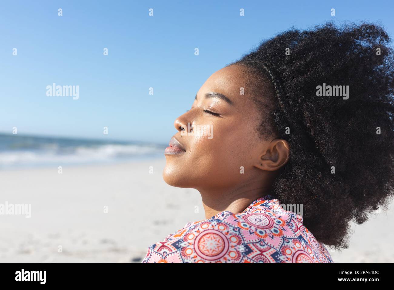 Rilassata donna afroamericana sorridente con gli occhi chiusi sulla spiaggia soleggiata in riva al mare Foto Stock