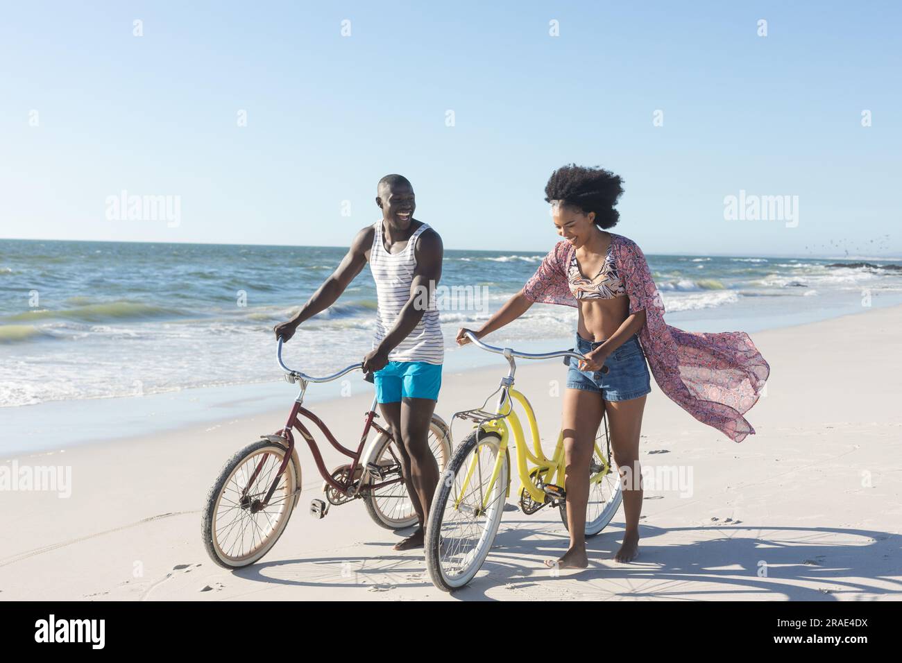Felice coppia afro-americana che cammina in bici sulla spiaggia soleggiata vicino al mare Foto Stock