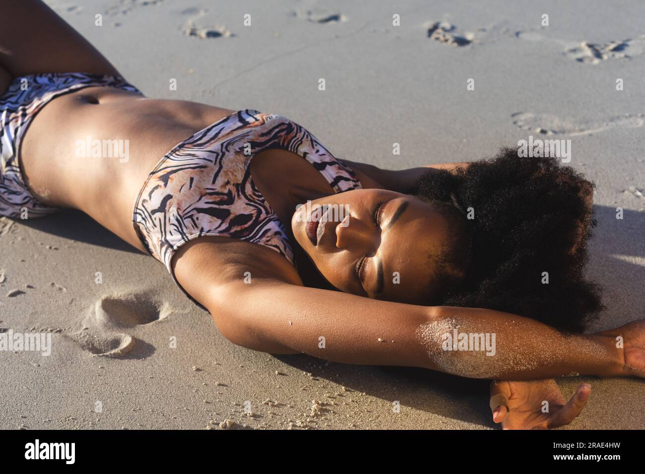 Felice donna afro-americana in bikini sdraiata sulla sabbia sulla spiaggia soleggiata Foto Stock