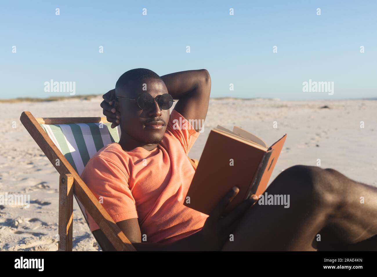 Felice uomo afroamericano con occhiali da sole seduto sulla sdraio leggendo un libro sulla spiaggia soleggiata Foto Stock