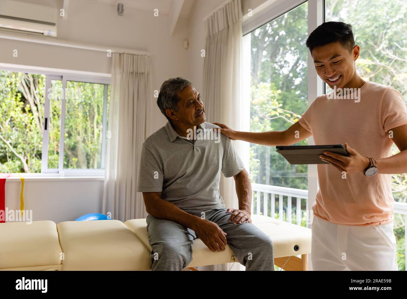 Fisioterapista maschile eterogeneo felice che esamina pazienti di sesso maschile anziani utilizzando la tavoletta Foto Stock