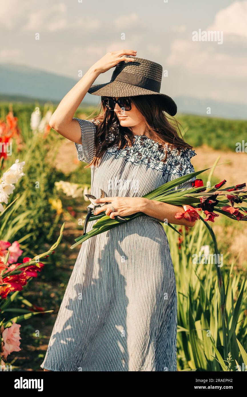 Splendida giovane donna che raccoglie fiori in un campo, indossa abiti estivi, cappello di paglia nero e occhiali da sole Foto Stock