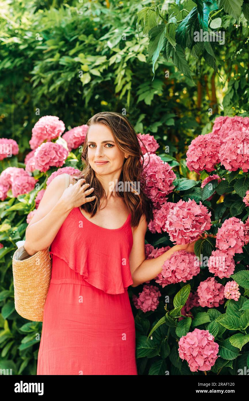 Ritratto all'aperto di una bella donna in posa con fiori di ortensia rosa, indossando un vestito rosso Foto Stock