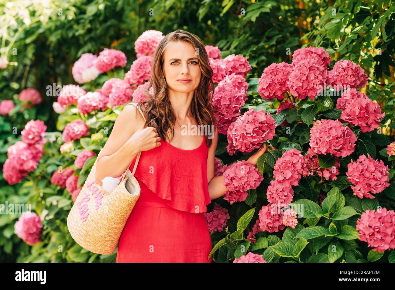 Ritratto all'aperto di una bella donna in posa con fiori di ortensia rosa, indossando un vestito rosso Foto Stock