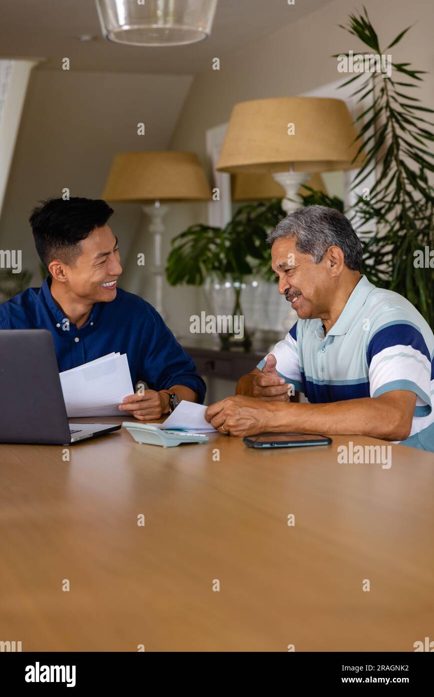 Consulente finanziario e uomo anziano felice e diversificato che discute di documenti cartacei e utilizza il computer portatile in sala da pranzo Foto Stock