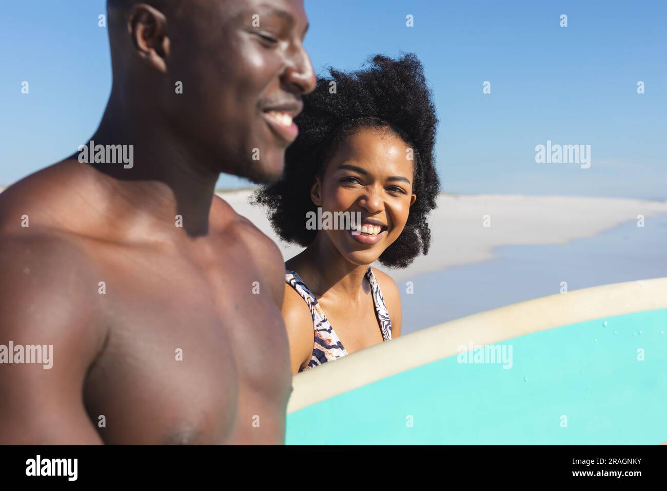 Coppia afroamericana felice e in forma che porta la tavola da surf sorridendo sulla spiaggia soleggiata in riva al mare Foto Stock