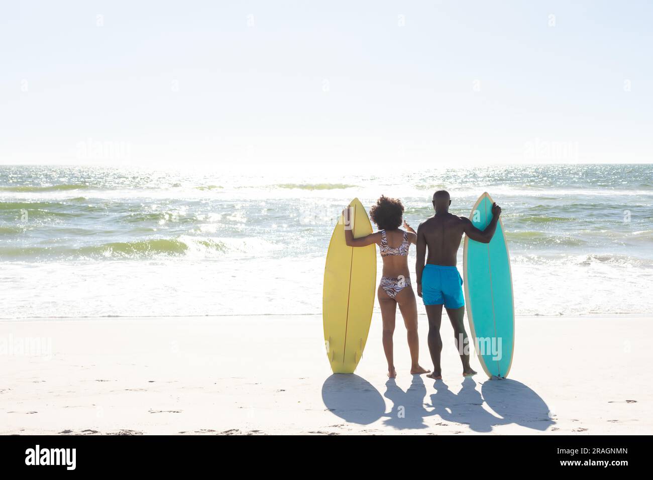 Vista posteriore di una coppia afroamericana con tavole da surf in piedi sulla spiaggia soleggiata di fronte al mare, spazio fotocopie Foto Stock