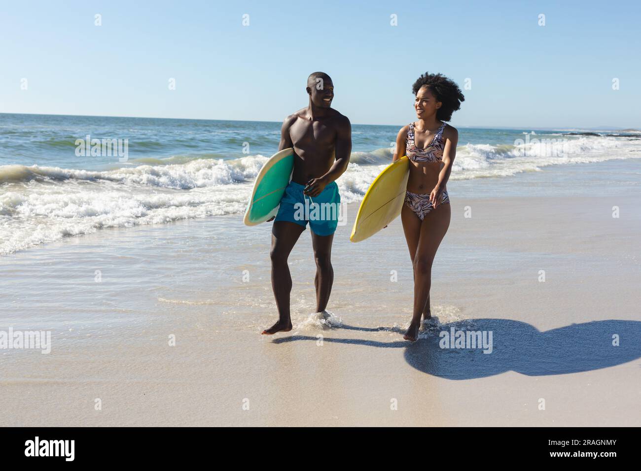 Coppia afroamericana felice e in forma che porta tavole da surf che cammina sulla spiaggia soleggiata in riva al mare Foto Stock