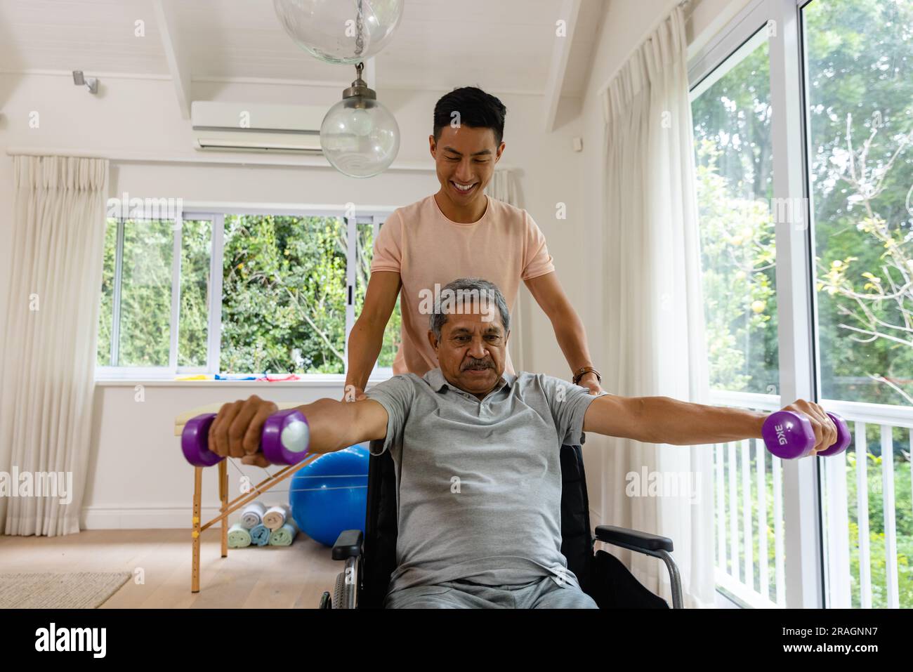Fisioterapista maschile felice eterogeneo che consiglia e paziente anziano in sedia a rotelle con manubri Foto Stock