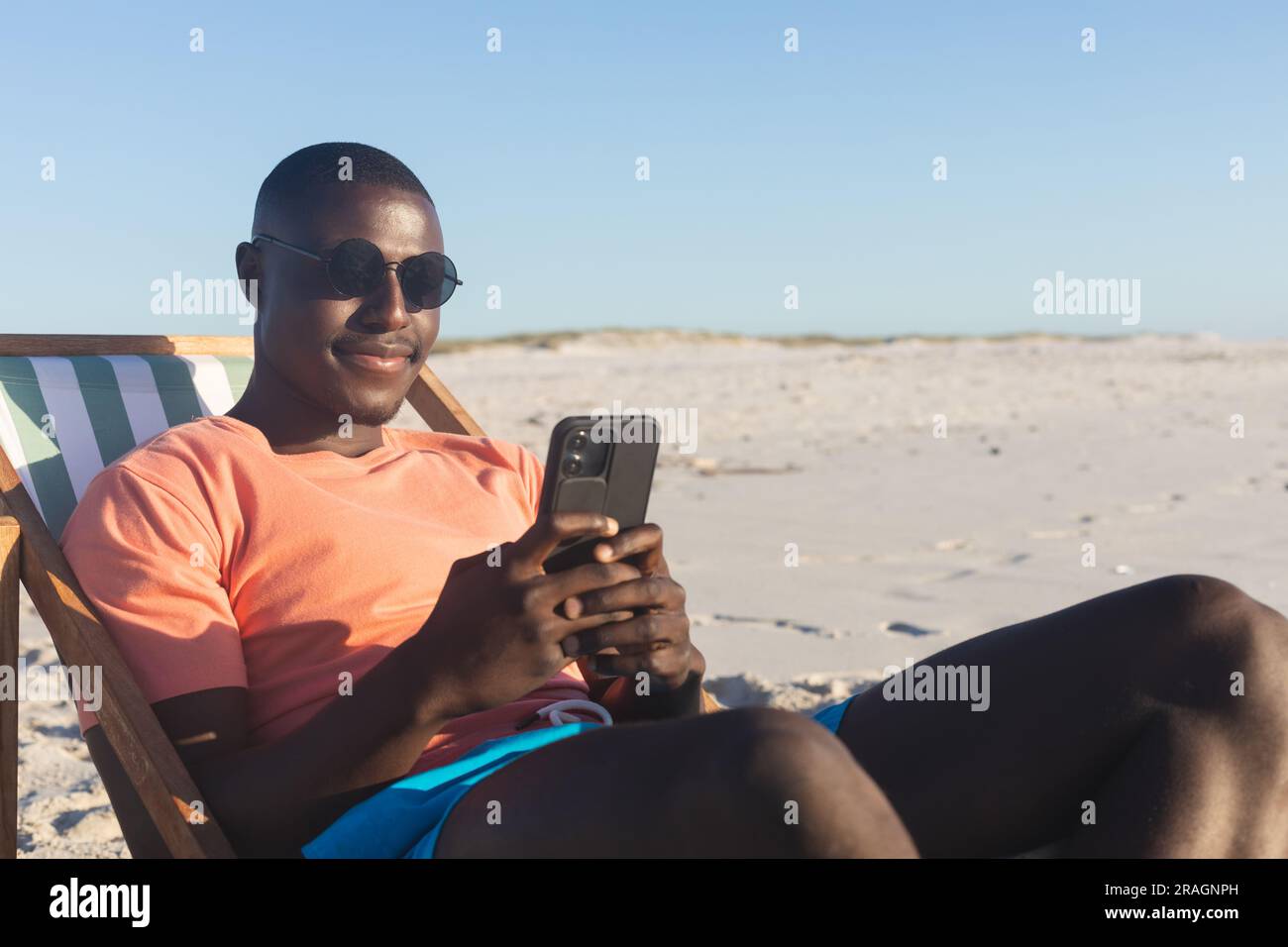 Felice uomo afro-americano con occhiali da sole seduto su una sdraio usando lo smartphone sulla spiaggia soleggiata Foto Stock