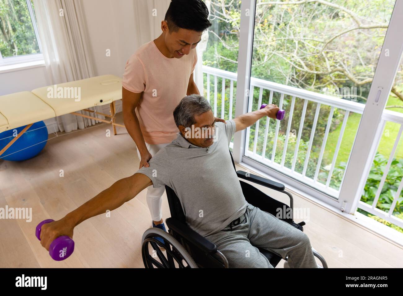 Fisioterapista maschile felice eterogeneo che consiglia e paziente anziano in sedia a rotelle con manubri Foto Stock