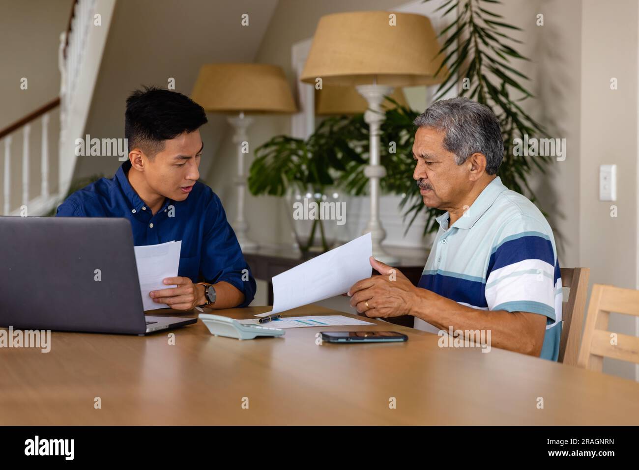 Consulente finanziario eterogeneo e uomo anziano che discute di documenti cartacei e utilizza il computer portatile in sala da pranzo Foto Stock