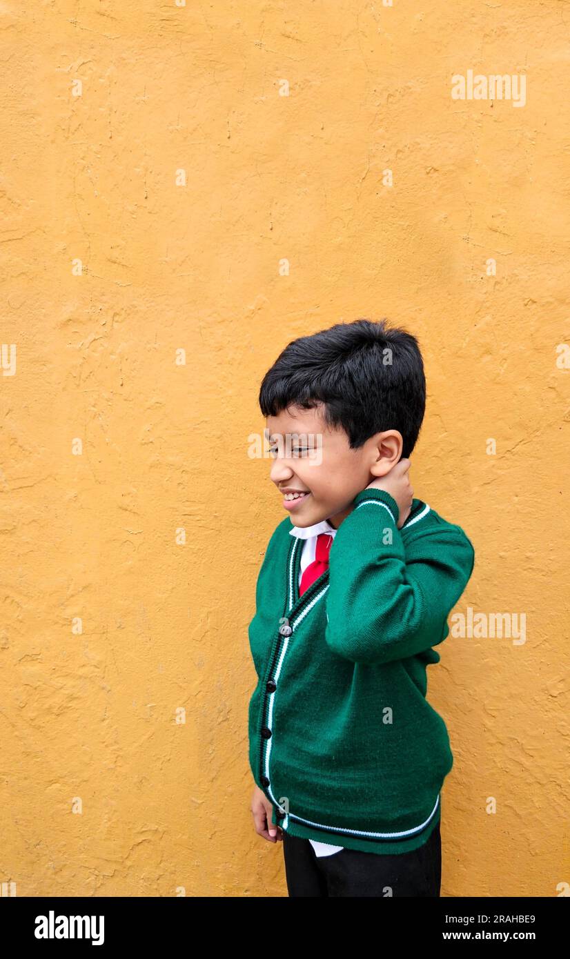 Un latinoamericano di 9 anni dalla pelle scura in uniforme scolastica soffre di dolore alla schiena e al collo a causa di un pesante zaino che soffre di tensione muscolare Foto Stock