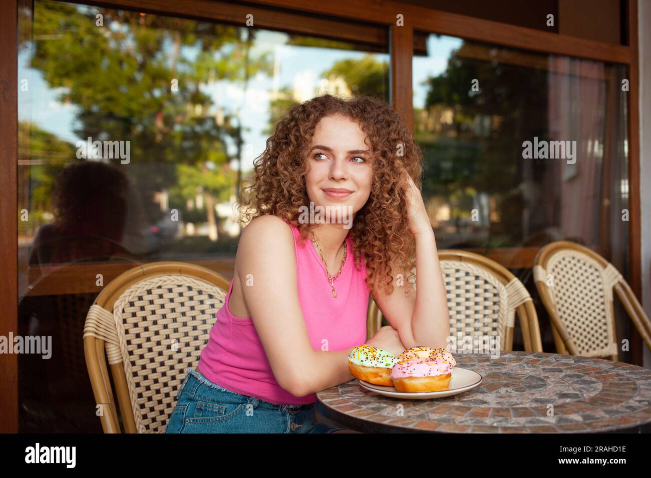 Ritratto di una bella giovane donna riccia nella caffetteria sulla terrazza estiva, deliziose ciambelle colorate, dolci dolci dolci. Pausa pranzo, sognante e felice. elegante c Foto Stock