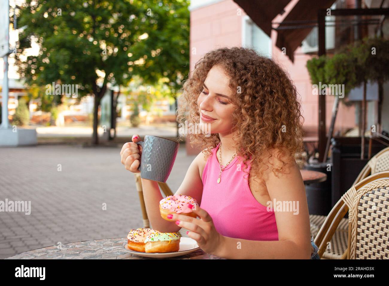 una giovane donna riccia siede al tavolo in una caffetteria sulla terrazza estiva. Deliziose ciambelle colorate, dolci dolci. Buon umore, sorridendo. Riposo, pausa snack Foto Stock