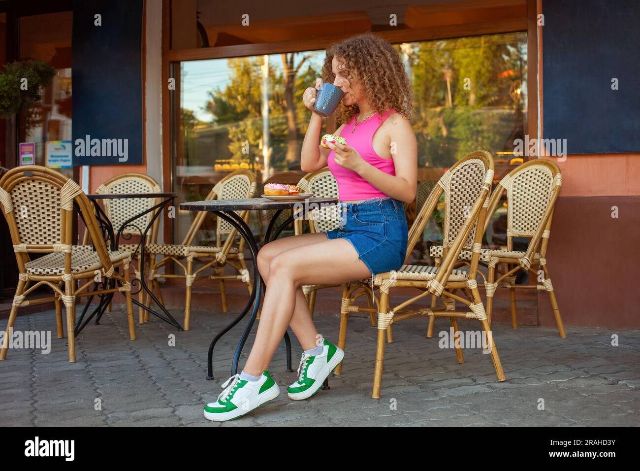 una giovane donna riccia siede al tavolo in una caffetteria sulla terrazza estiva. Deliziose ciambelle colorate, dolci dolci. Buon umore, sorridendo. Riposo, pausa snack Foto Stock