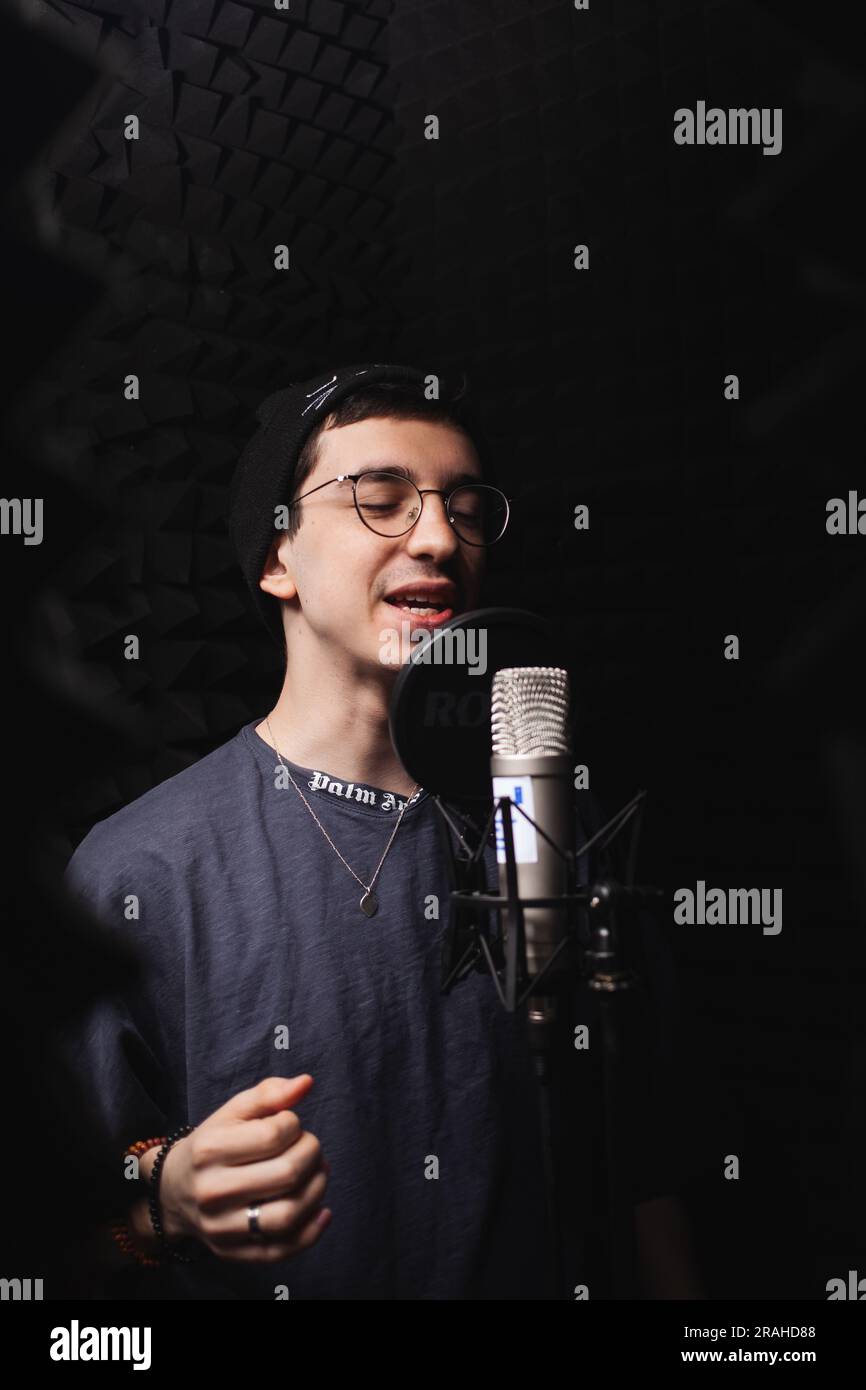 l'uomo canta nel microfono, nello studio di registrazione professionale, nelle cuffie. Pareti nere morbide, isolamento acustico. cantante registra la sua canzone. uomo con gli occhiali Foto Stock