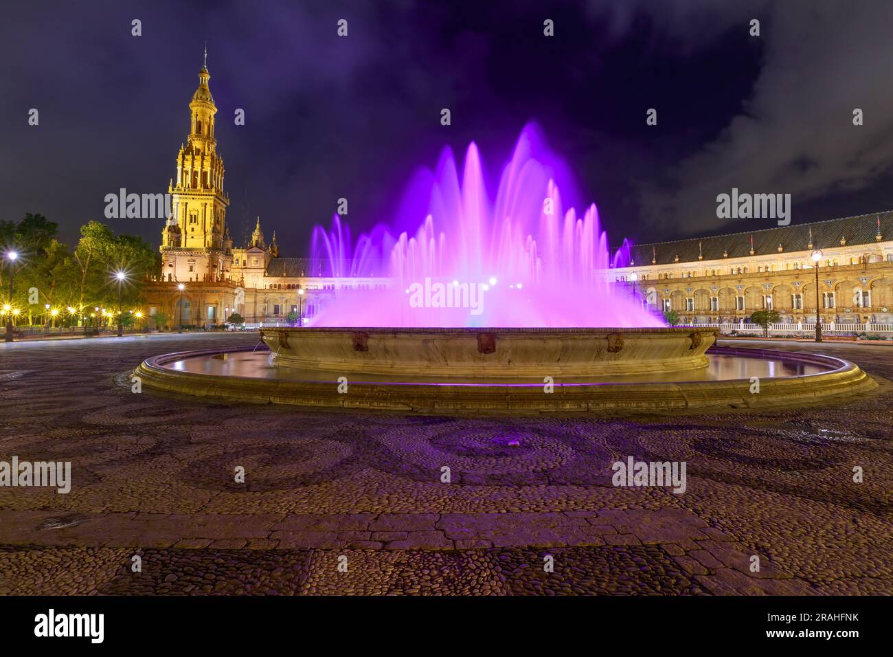 Una luminosa fontana nella Plaza de Espana di Siviglia si anima di notte, con le sue acque violette in aggraziato movimento. Accattivanti spettatori in questa incantevole atmosfera Foto Stock