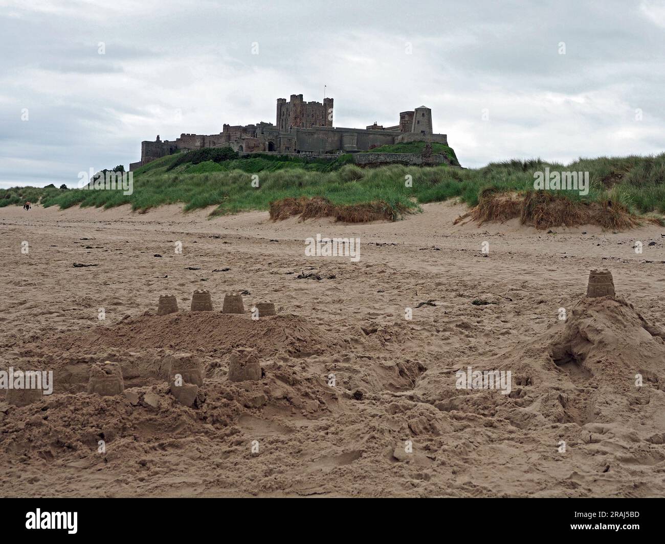Possente fortificazione del castello di Bamburgh che torreggia sulle dune di sabbia e sulla spiaggia del Mare del Nord con pallide imitazioni di castelli di sabbia in Northumbria, Inghilterra, Regno Unito Foto Stock