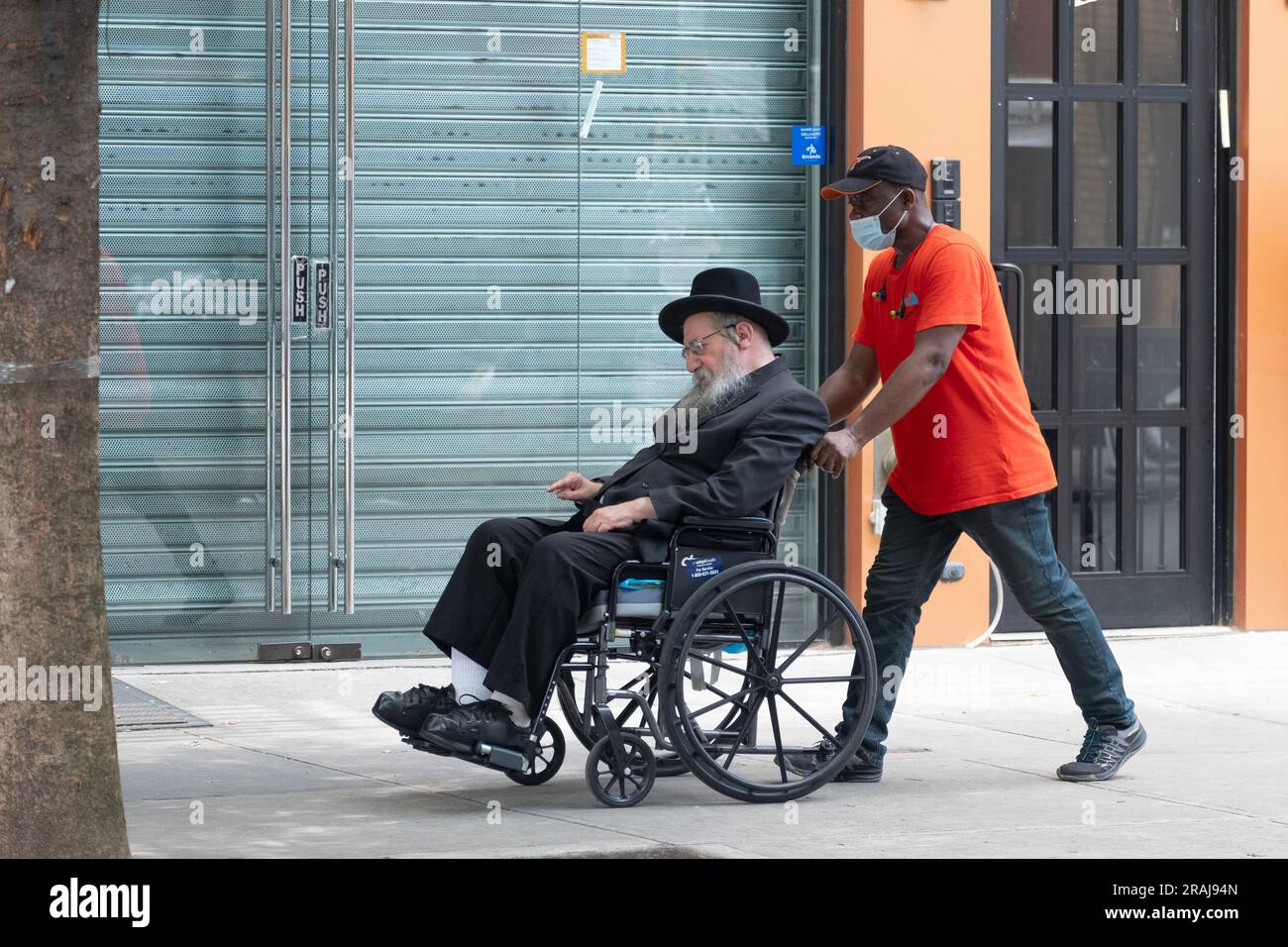 Un anziano ebreo chassidico sulla sua sedia a rotelle assistito dal suo assistente. In una strada tranquilla a Brooklyn, New York. Foto Stock