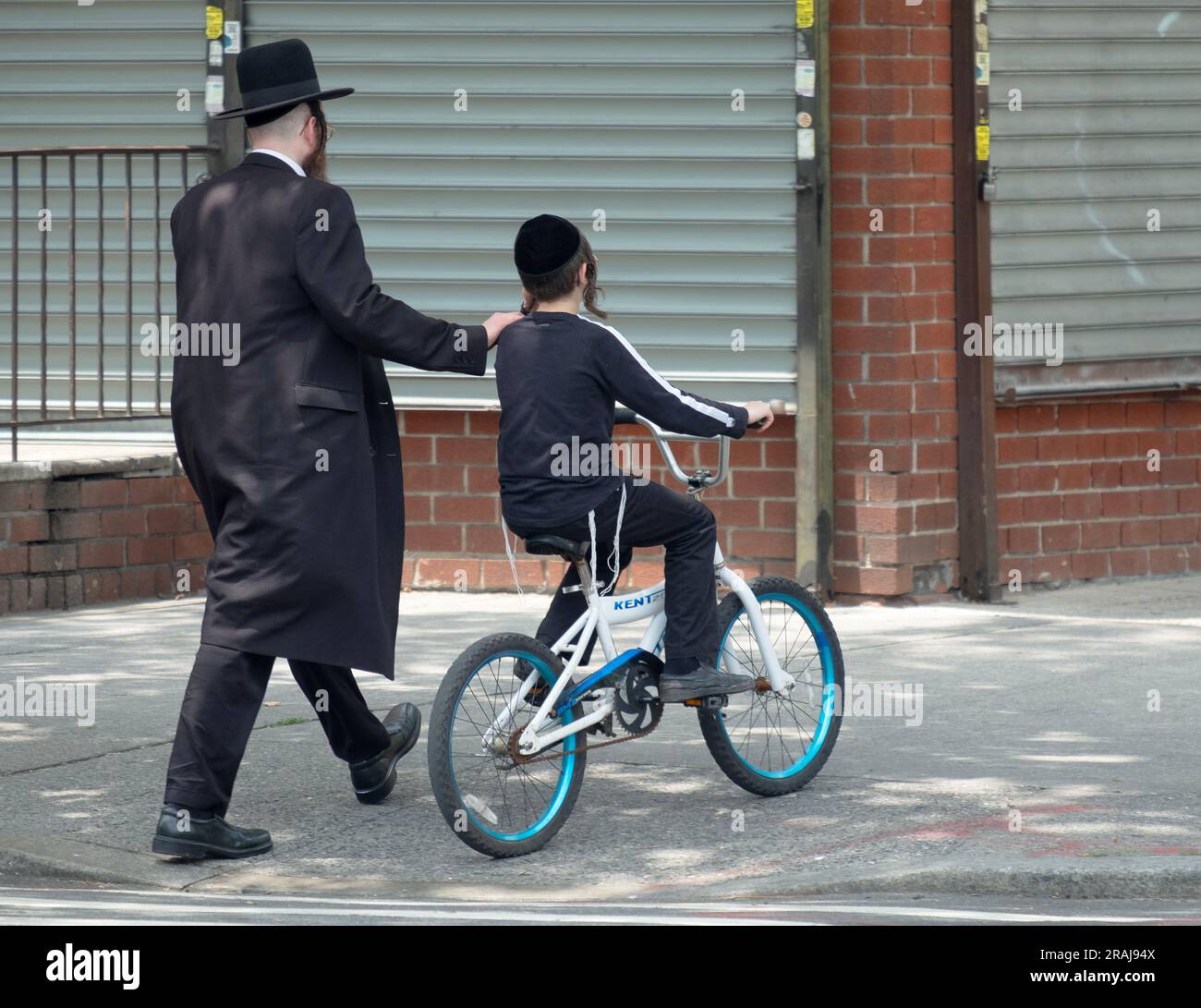 Un padre chassidico presta supporto al figlio mentre cammina/pedala su una strada trafficata di Brooklyn, New York Foto Stock