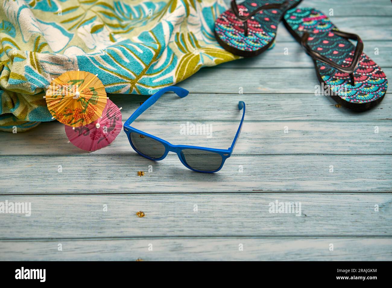 Occhiali da sole blu con due piccoli ombrelli arancioni e rosa, un asciugamano con stampa tropicale e infradito, sopra un tavolo di legno. Foto Stock