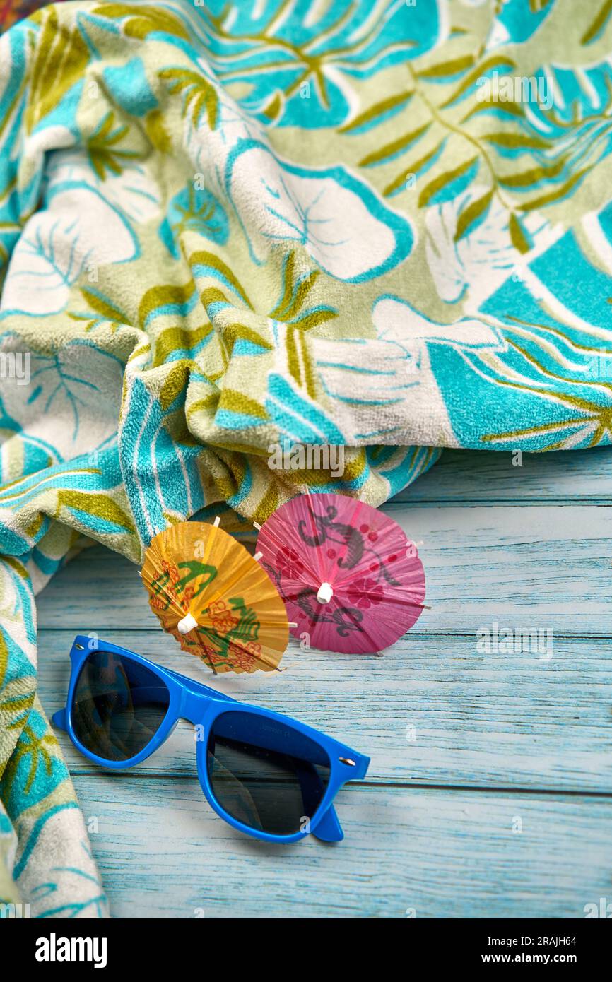 Occhiali da sole blu, piccoli ombrelli arancioni e rosa e telo da spiaggia con stampa tropicale su un tavolo di legno. Foto Stock