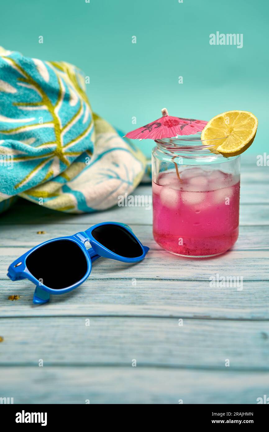 Bevi un rosa rinfrescato con un ombrello e una fetta di limone, occhiali da sole blu e un telo da spiaggia con un motivo tropicale su un tavolo di legno e un bac blu Foto Stock