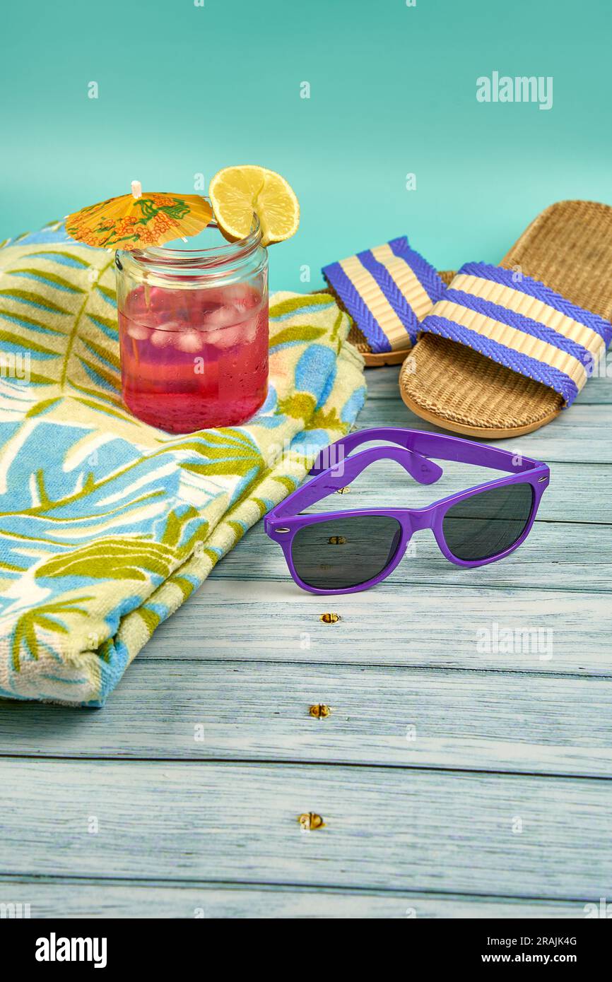 Rinfrescante bevanda rosa con un ombrello e una fetta di limone, occhiali da sole viola, asciugamano e sandali da spiaggia, su un tavolo di legno e uno sfondo verde. Foto Stock