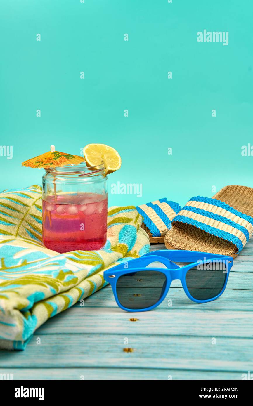 Rinfrescante bevanda di colore rosa con un ombrello e una fetta di limone, occhiali da sole blu, asciugamano e sandali da spiaggia, su un tavolo di legno e uno sfondo verde Foto Stock