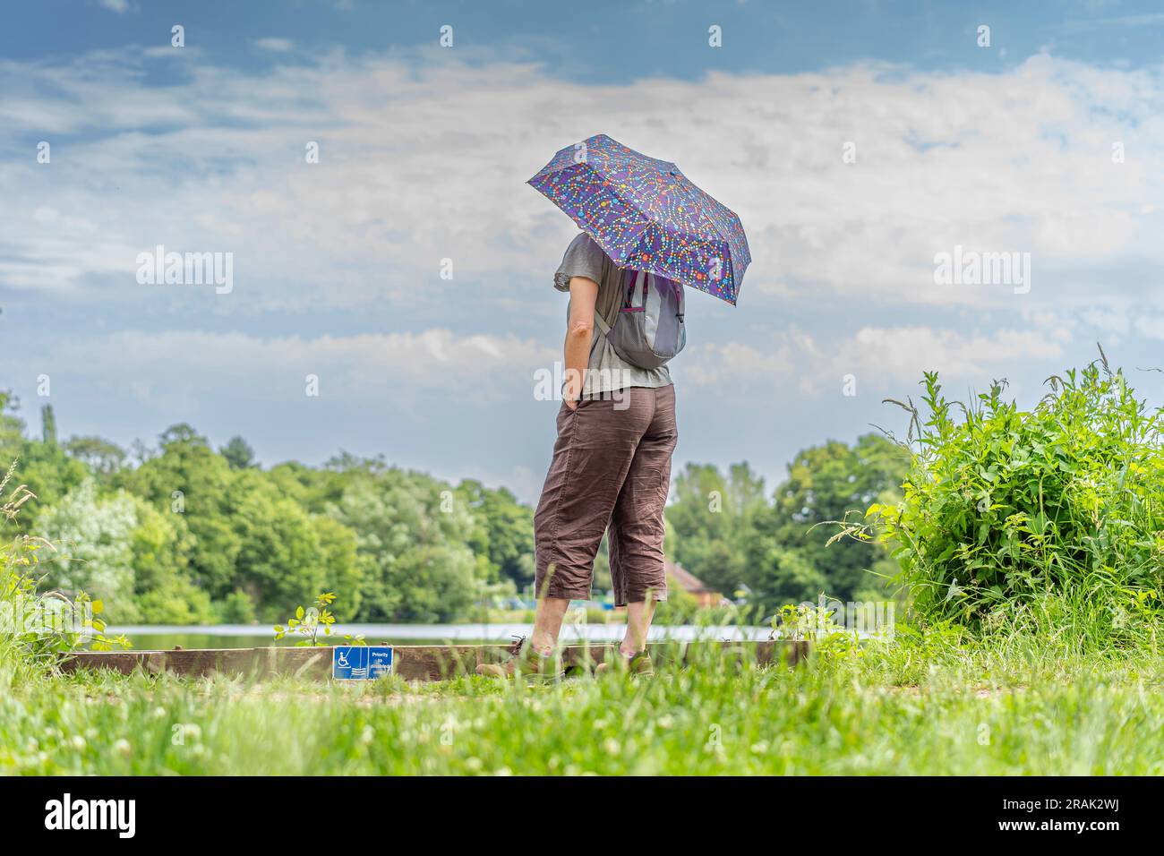Vista posteriore pov bassa di una donna con una brolla solare in piedi di fronte a una piscina in una giornata di sole. Foto Stock