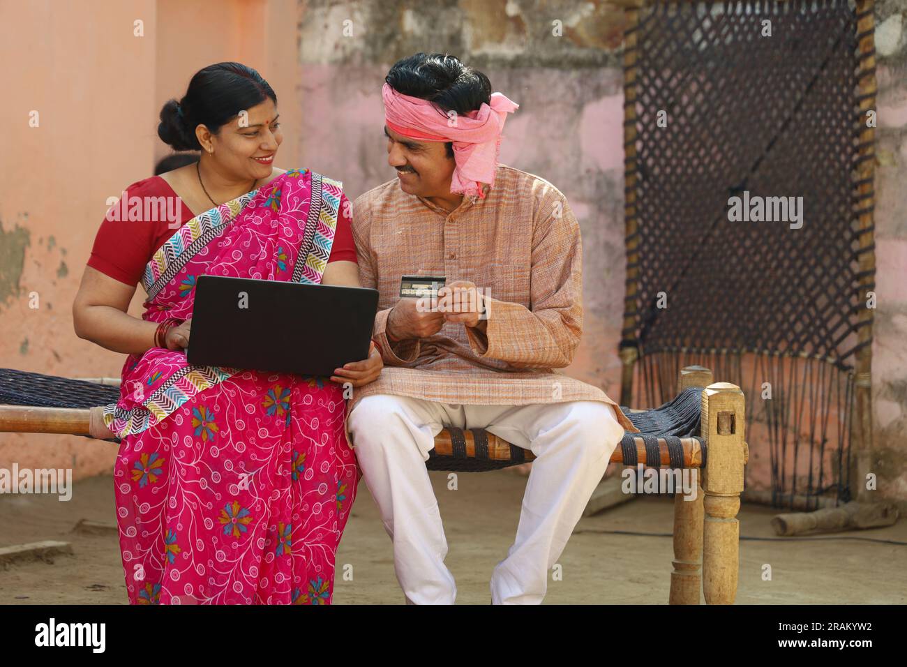 Una felice famiglia di villaggi indiani che usa il computer portatile seduto fuori casa. Marito e moglie sono felici di effettuare il pagamento online con carta di credito in mano Foto Stock