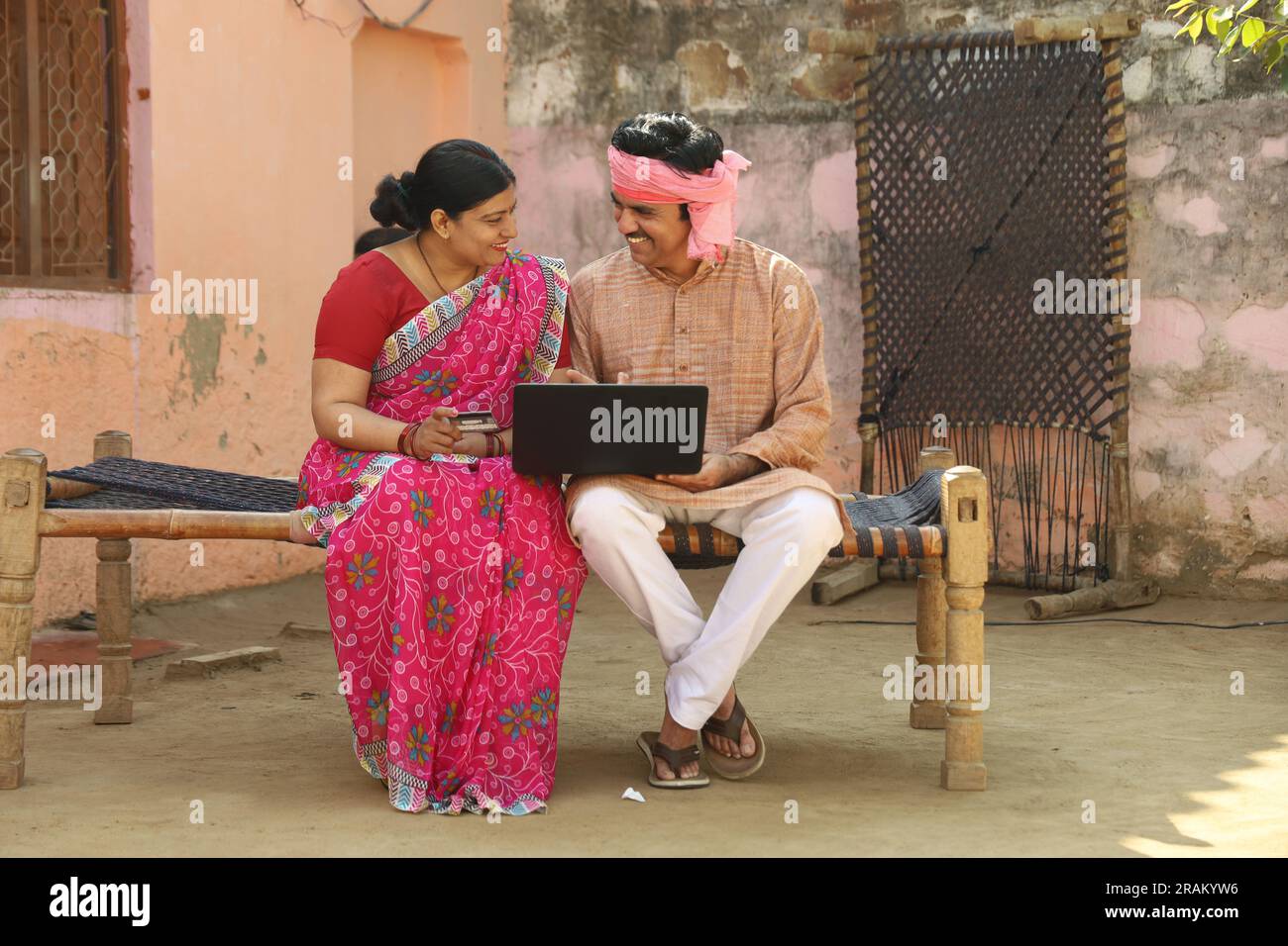 Una felice famiglia di villaggi indiani che usa il computer portatile seduto fuori casa. Marito e moglie sono felici di effettuare il pagamento online con carta di credito in mano Foto Stock