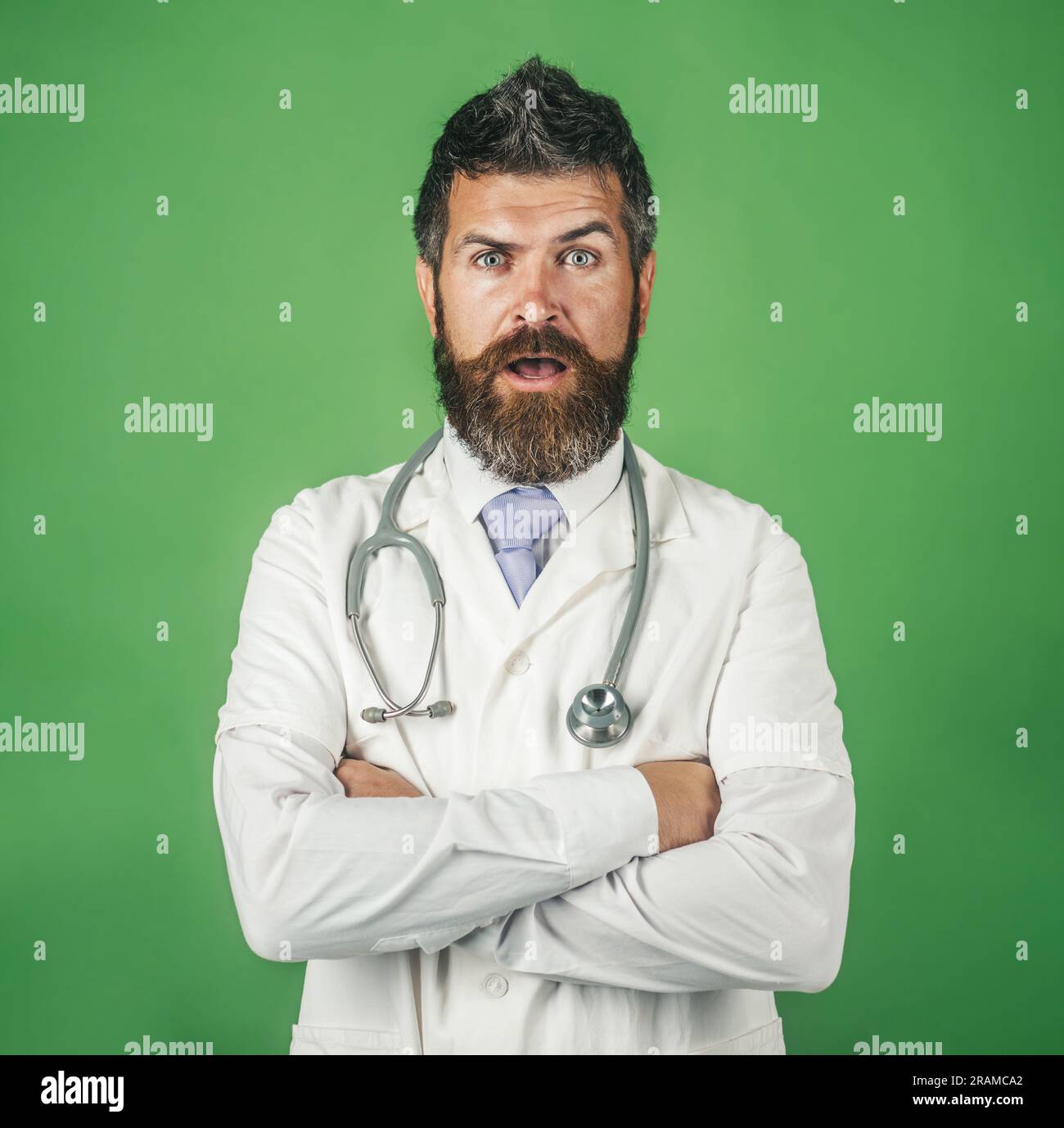 Stupito medico maschile in abito medico con stetoscopio sul collo. Medico professionista in abito medico con braccia piegate. Medico barbuto in abbigliamento da lavoro con Foto Stock