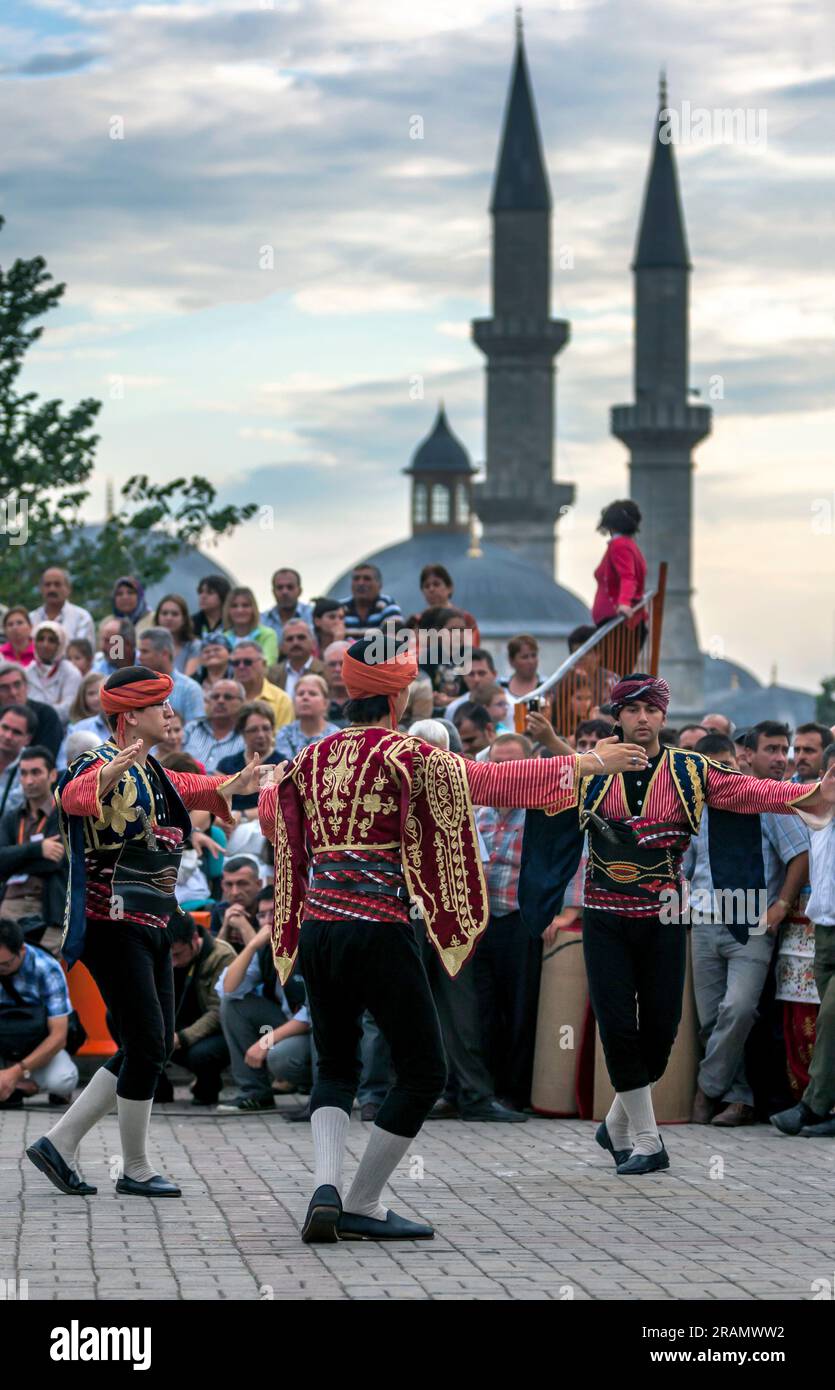 I ballerini turchi vestiti con costumi dell'epoca ottomana si esibiscono a Edirne in Turkiye. Sullo sfondo si trovano i minareti di Eski Ulu Camii. Foto Stock