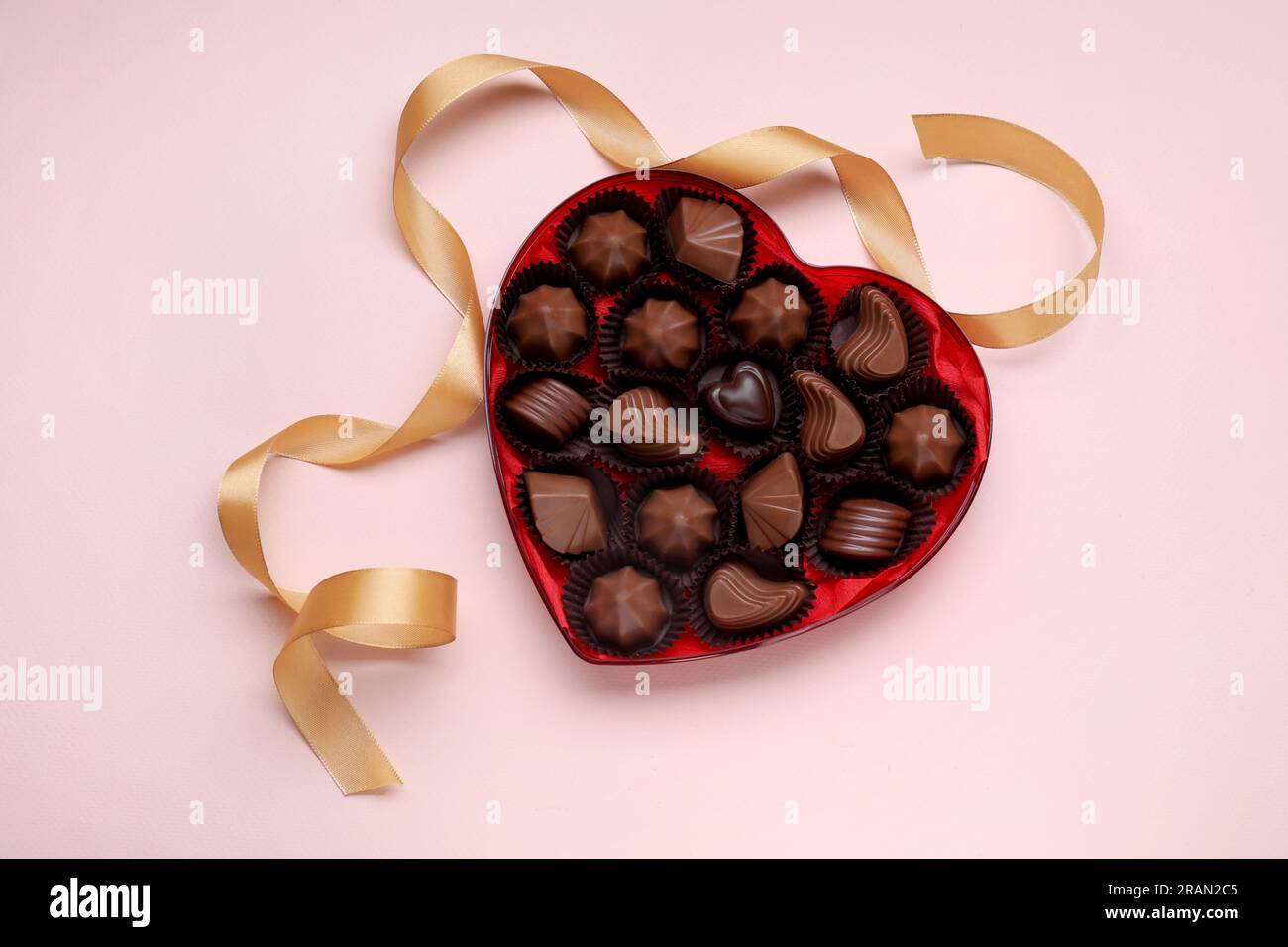 Scatola a forma di cuore con deliziose caramelle al cioccolato e nastro su sfondo rosa pallido, piatto Foto Stock