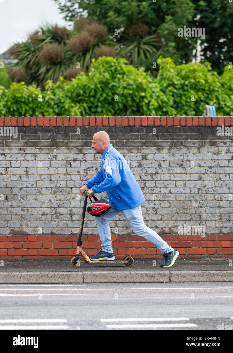 Uomo che guida su uno scooter per bambini a propulsione manuale lungo un marciapiede in Inghilterra, Regno Unito. Foto Stock