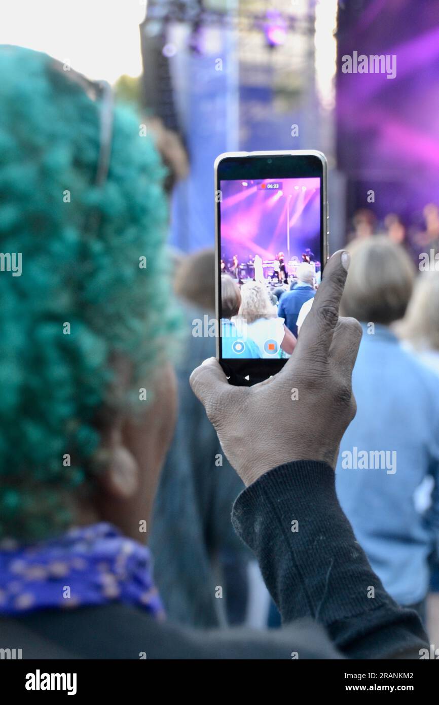 Signora anziana con i capelli verdi che filma un concerto rock in un festival con l'immagine del palco e della band sullo schermo dello smartphone Foto Stock